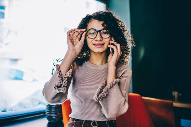 Half length portrait of smiling hipster girl in stylish spectacles calling to friend to discuss time of meeting, young positive woman looking at camera during telephone conversation at campus - Foto, afbeelding