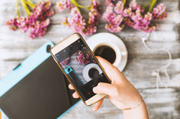 instagram photography blogging workshop concept, hand holding phone and taking photo of stylish flowers, cup of coffee and graphic tablet on grey wood rustic background.space for text - Foto, Bild