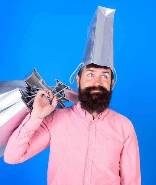 Man with beard and mustache carries shopping bags, blue background. Hipster on deamy face with bag on head is addicted shopaholic. Guy shopping on sales season with discounts. Shopping concept - 写真・画像