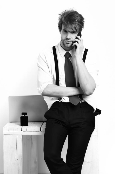 Bearded man, short beard. Caucasian stylish manager with moustache in red tie, shirt with suspenders sitting on table and speaking be cell phone in studio on white background - Photo, image