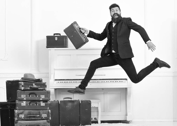 Macho attractive, elegant on cheerful face carries vintage suitcases, jumping. Long awaited vacation concept. Man with beard and mustache in suit carries luggage, luxury white interior background - Фото, изображение