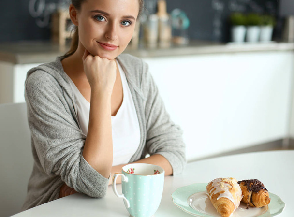 Woman drinking tea with sweet croissant at the kitchen table - 写真・画像