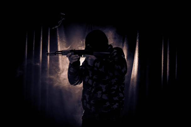 Σιλουέτα του ανθρώπου με την επίθεση τουφέκι έτοιμος να επιτεθεί σε σκούρο φόντο τονισμένο ομίχλη ή επικίνδυνο ληστή σε μαύρο, φορώντας κουκούλα και εκμετάλλευση όπλο στο χέρι. Γυρίσματα τρομοκράτης με το όπλο θέμα ντεκόρ - Φωτογραφία, εικόνα