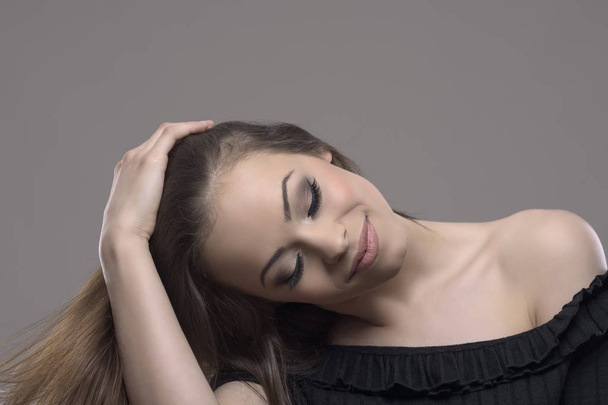 entspannte hübsche junge Frau brünette Berührung der Haare mit geschlossenen Augen vor grauem Studiohintergrund mit Copyspace. - Foto, Bild
