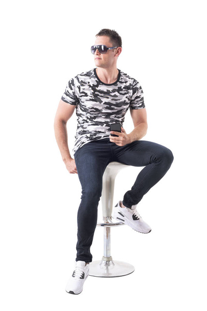 Confident handsome man in camo shirt with sunglasses sitting on bar stool looking away. Full body isolated on white background. - Φωτογραφία, εικόνα