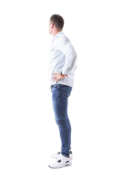 Vista lateral do jovem em camisa azul clara e jeans com as mãos nos quadris olhando para trás. Corpo inteiro isolado em fundo branco
. - Foto, Imagem