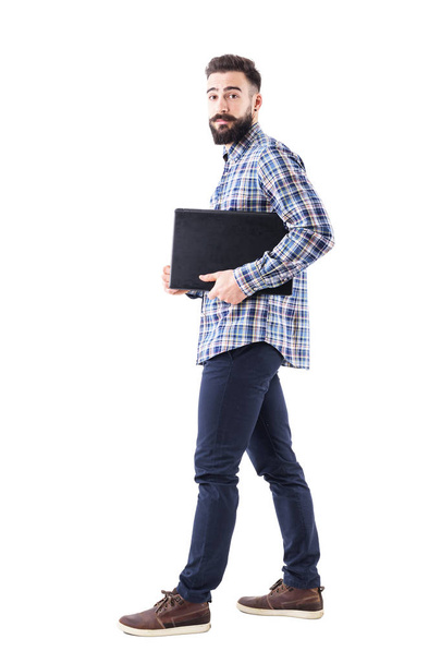Stylish modern bearded business man holding laptop under arm walking. Side view. Full body isolated on white background. - Photo, image