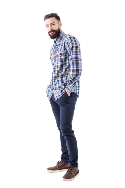 Happy young bearded man in checked shirt with hands in pockets smiling and looking at camera. Full body isolated on white background. - Fotografie, Obrázek