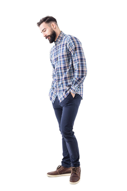 Shy handsome bearded young man in plaid shirt with hands in pockets smiling and looking down. Full body isolated on white background. - 写真・画像