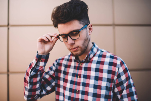 Knappe jongeman in trendy shirt en brillen poseren op muur achtergrond buitenshuis, slimme mannelijke student gekleed in stijlvolle outfit denken over onderwijs nieuwe bril aan te raken voor de bescherming van de visie - Foto, afbeelding
