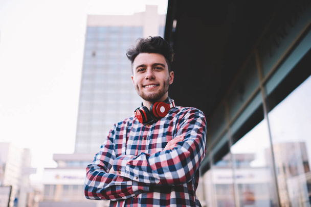Half length portrait of confident hipster guy standing with crossed arms outdoors, handsome positive young man in stylish shirt and with modern red headphones looking at camera posting on urban setting - Foto, afbeelding