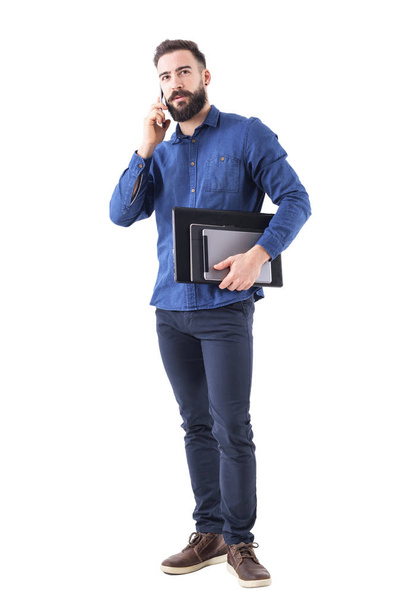 Confident successful young ceo talking on the phone looking up and holding tablet and laptop under arm. Full body isolated on white background. - Photo, Image