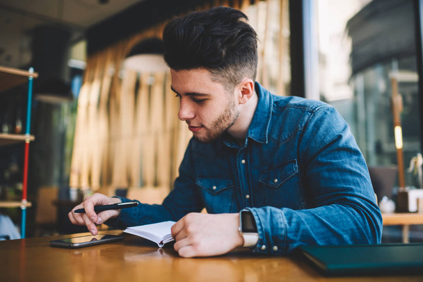 Young male student using smartphone for browsing information and making notes while sitting at cafe interior, concentrated hipster guy writing plans in notepad using organizing apps on cellular - Fotoğraf, Görsel