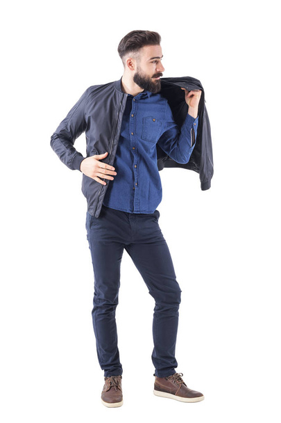 Bearded handsome man putting on bomber jacket and looking away. Full body length portrait isolated on white studio background. - Photo, Image