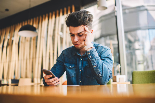 Stressed male feeling headache getting bill on smartphone email sitting at table in cafeteria, disappointed hipster guy reading bad news in income message on telephone having pain about trouble - Photo, Image