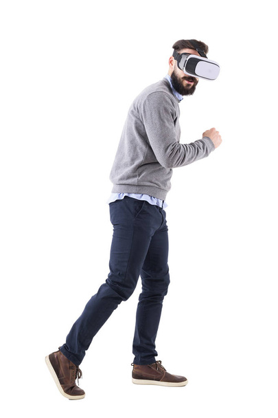 Side view of young bearded smart casual man with vr glasses running astonished and looking at camera. Full body isolated on white background. - Foto, imagen