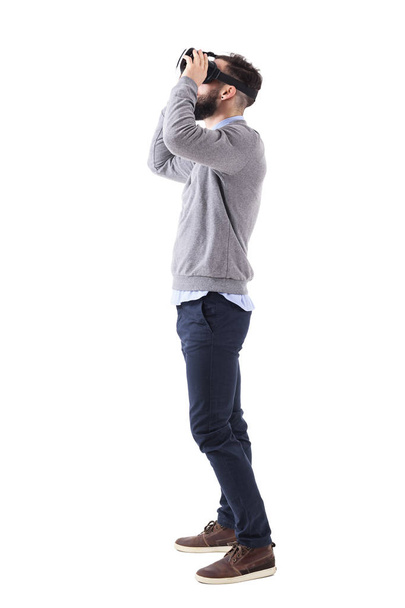 Vista de perfil de un joven que tiene experiencia en realidad virtual sosteniendo gafas y mirando hacia arriba. Cuerpo completo aislado sobre fondo blanco
. - Foto, Imagen