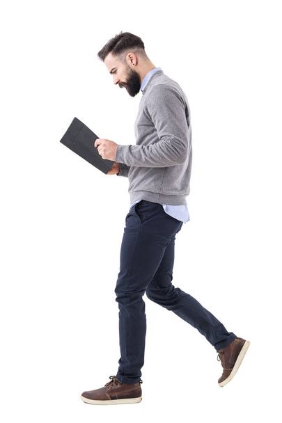 Vue du profil d'un jeune homme d'affaires barbu marchant en lisant un cahier ou un planificateur. Portrait intégral isolé sur fond de studio blanc
. - Photo, image