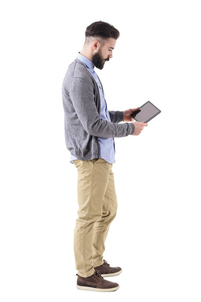 Elegante hombre de negocios hipster sosteniendo y viendo tablet pad ordenador. Vista lateral. Retrato de cuerpo entero aislado sobre fondo blanco de estudio
. - Foto, Imagen