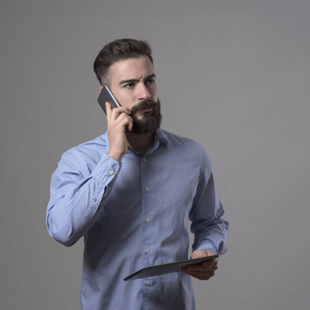 Jistý mladý vousatý obchodní muž mluví na mobil drží tabletový počítač a hledat dál přes šedý studio pozadí. - Fotografie, Obrázek