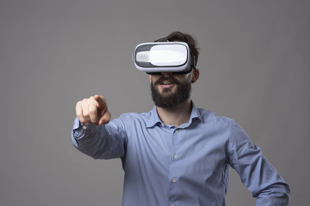 Amazed young bearded adult business man with vr headset touching virtual digital touch screen over gray studio background. - Photo, Image