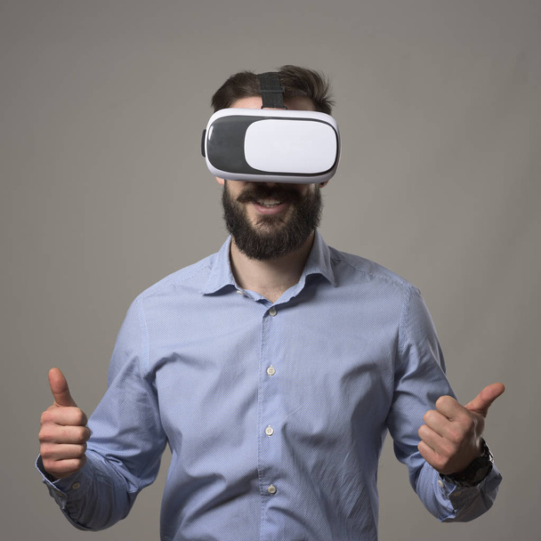 Young adult bearded smart casual business man wearing vr headset smiling with thumbs up against gray studio background. - Photo, Image