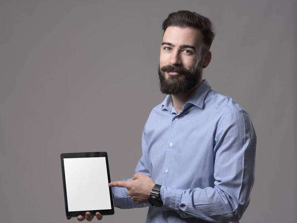 Jovem sorridente amigável homem de negócios apontando dedo e publicidade vazio tablet monitor de computador em branco sobre fundo estúdio cinza
. - Foto, Imagem