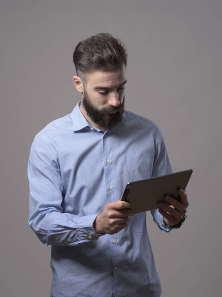 Standing young adult bearded business man holding and watching at tablet computer over gray studio background. - Fotoğraf, Görsel