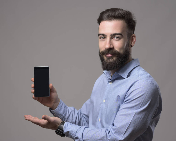 junger bärtiger lächelnder positiver Geschäftsmann mit leerem Smartphone-Display vor grauem Hintergrund.  - Foto, Bild