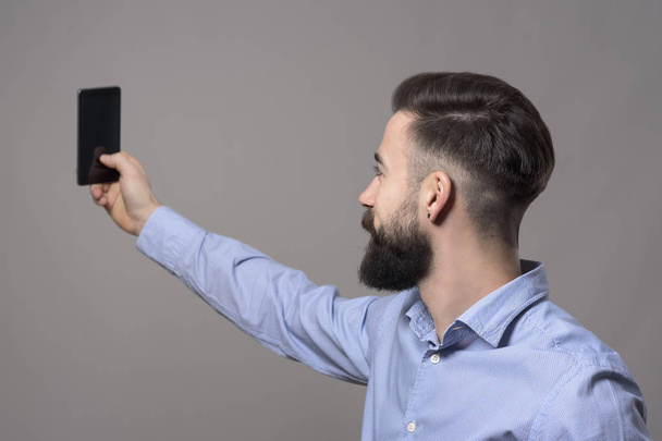 Vue du profil de jeune homme d'affaires barbu moderne prenant des photos selfie avec smartphone sur fond de studio gris
. - Photo, image