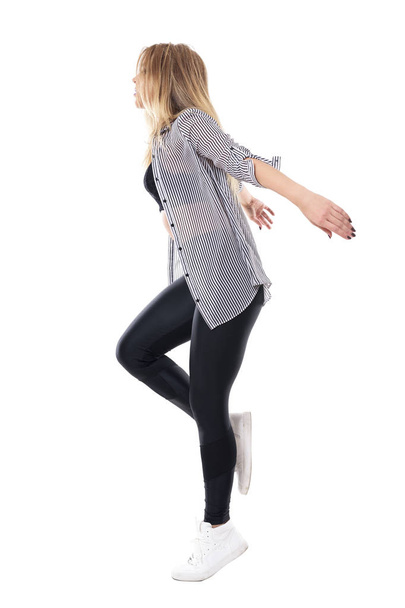 Πλαϊνή όψη, χαριτωμένη γυναίκα τζαζ χορευτής χορεύει και άλματα στο ένα πόδι με τα χέρια πίσω από. Γεμάτο σώμα μήκος πορτραίτο απομονώνονται σε φόντο λευκό στούντιο. - Φωτογραφία, εικόνα