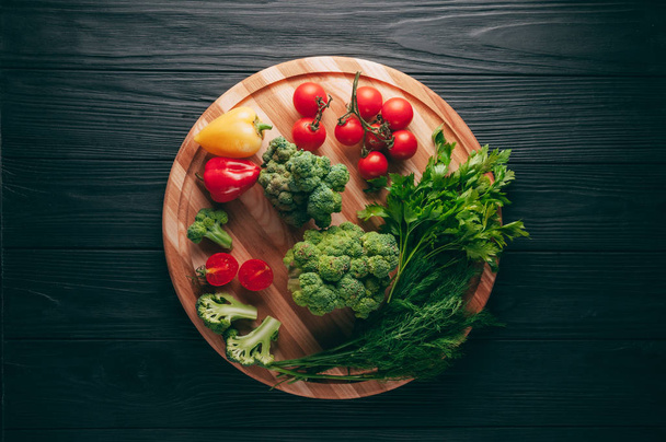 Fresh vegetables: cherry tomatoes, broccoli, pepper, dill, parsley on a round wooden board, on a dark wooden table. Recipe. Ingredients. Dietary food. Place under the text. View from above. - 写真・画像