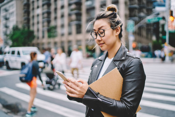 Young attractive female blogger in glasses for eye protection sharing news with followers via smartphone application.Gorgeous hipster girl strolling at urban setting with gadget during leisure time - Fotografie, Obrázek
