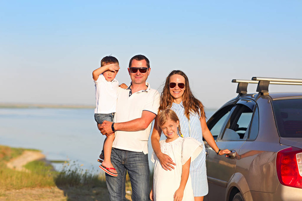 Deniz arka planı olan arabanın yanında iki çocuklu aile gülümseyen mutlu. Arabanın yanında plajda iki çocuklu gülümseyen bir aile portresi. Tatil ve seyahat kavramı - Fotoğraf, Görsel