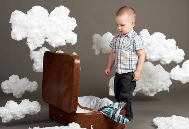 το αγόρι κάθεται σε μια βαλίτσα και πηγαίνει σε ένα ταξίδι, σύννεφα, βαμβάκι ως διακόσμηση, γκρι φόντο - Φωτογραφία, εικόνα