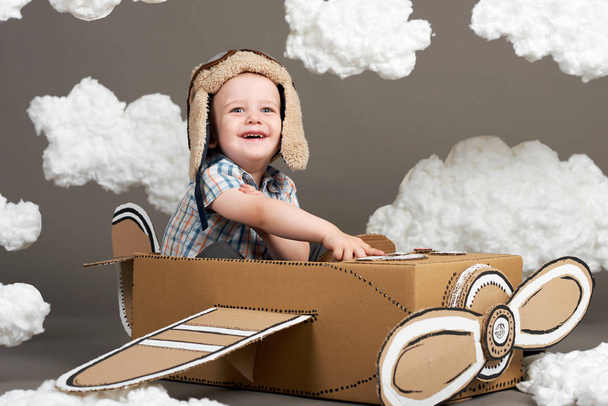 the boy plays in an airplane made of cardboard box and dreams of becoming a pilot, clouds from cotton wool on a gray background, retro style - Photo, Image
