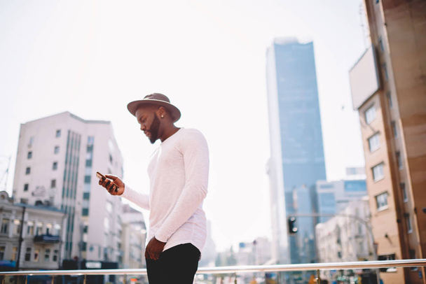 Young african american trendy dressed guy checking notification on cellphone standing on urban setting background, dark skinned male hipster guy using app on smartphone  and internet in roaming - Fotografie, Obrázek