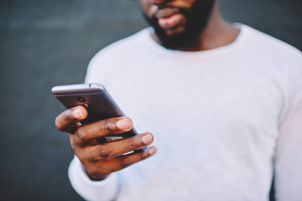 Cropped image of african american man holding modern smartphone dialing number, dark skinned male in white shirt using mobile phone for chatting in social networks via 4G internet connection outdoors - Photo, Image