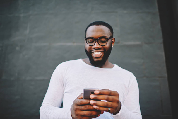Vrolijke Afro-Amerikaanse hipster kerel lezing informatie van cellulaire doorbladerde web-pagina permanent buitenshuis, glimlachend van donkere gevilde man blij met nieuw inkomen-bericht met roaming internet krijgen - Foto, afbeelding