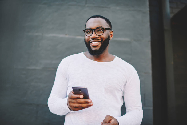 Cheerful african american male in spectacles using mobile phone outdoors looking waya and smiling, positive dark skinned hipster guy in white shirt with copy space for brandname holding cellular - Fotografie, Obrázek