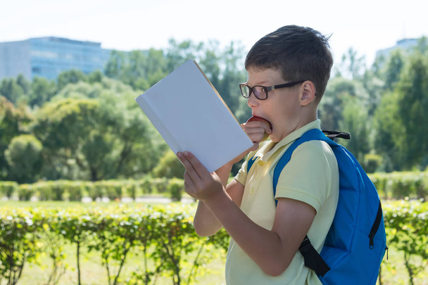 un niño con un maletín leyendo un libro y mordiendo una manzana en la calle
 - Foto, imagen