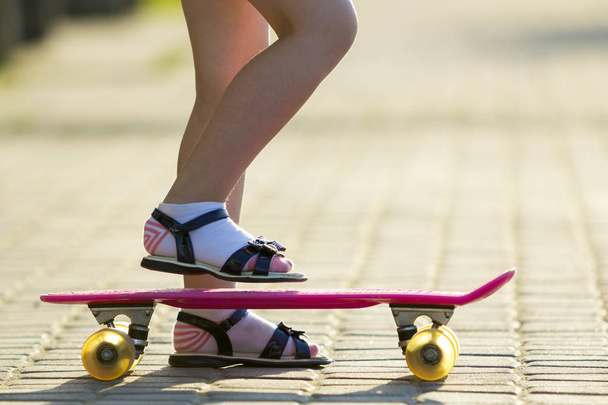 Child slim legs in white socks and black sandals on plastic pink skateboard on bright sunny summer blurred copy space pavement background. Outdoors activities and healthy lifestyle concept. - Photo, Image