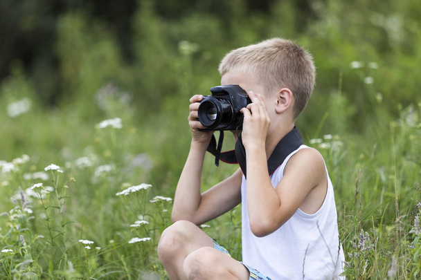 Profile close-up portrait of young blond cute handsome child boy with camera taking pictures outdoors on bright sunny spring or summer day on blurred light green grassy copy space background. - Φωτογραφία, εικόνα