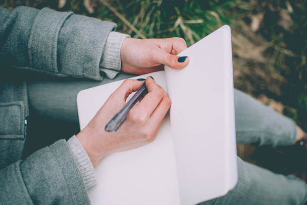 Roodharige meisje zittend op het gras van de groene lente haar gedachten in notitieblok opschrijven. Bleke handen en blanco papier van notebook. Concept (idee) van denken, dromen, motivatie, plannen, studeren. - Foto, afbeelding