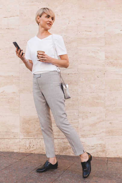 Full length image of stylish blond woman wearing white t-shirt using cell phone while standing against beige wall outdoor in summer and looking aside at copyspace with takeaway coffee - Valokuva, kuva