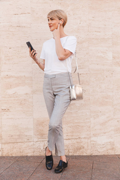 Full length image of stylish blond woman 30s wearing white t-shirt using mobile phone while standing against beige wall outdoor and touching bluetooth earphone - Fotó, kép