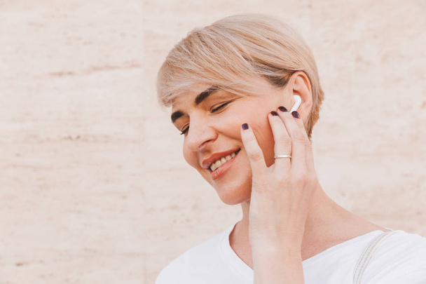 Photo closeup of happy blond woman wearing white t-shirt smiling and touching bluetooth wireless earphone while standing against beige wall outdoor - Foto, Imagen