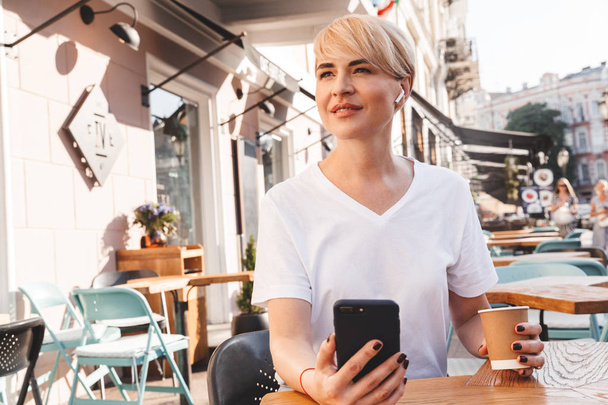 Image of pretty adult woman wearing white t-shirt using cell phone and wireless earphone while sitting in cafe outside in summer and drinking coffee from paper cup - 写真・画像