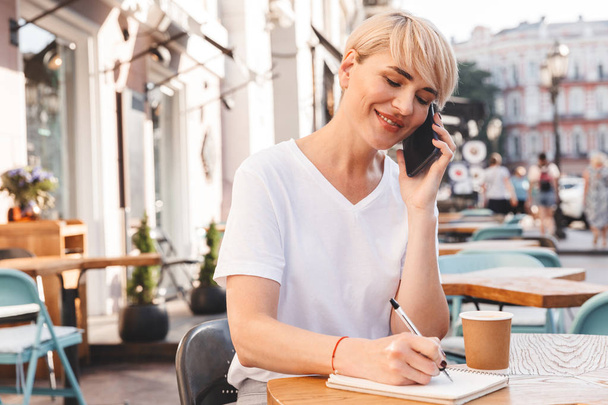 European adult woman wearing white t-shirt sitting in summer cafe outdoor while writing down in notebook and talking on smartphone - Foto, Imagen