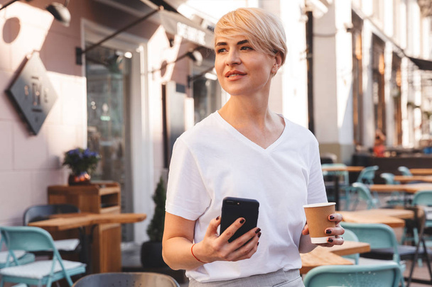 Portrait of satisfied mature woman wearing white t-shirt using smartphone while resting in cafe outdoor in summer and drinking coffee from paper cup - Foto, Bild
